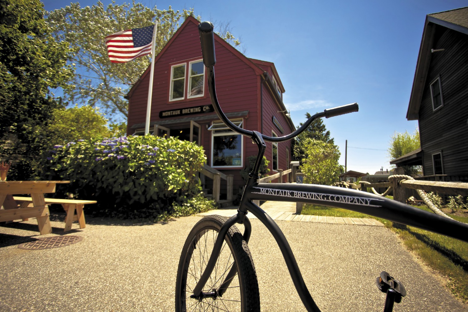 Photo of Montauk Brewing Company. There is a vintage black bike with the words "Montauk Brewing Company" in white across the frame sitting in front of the camera and the large dark red wood building of Montauk Brewing Company is in the background.