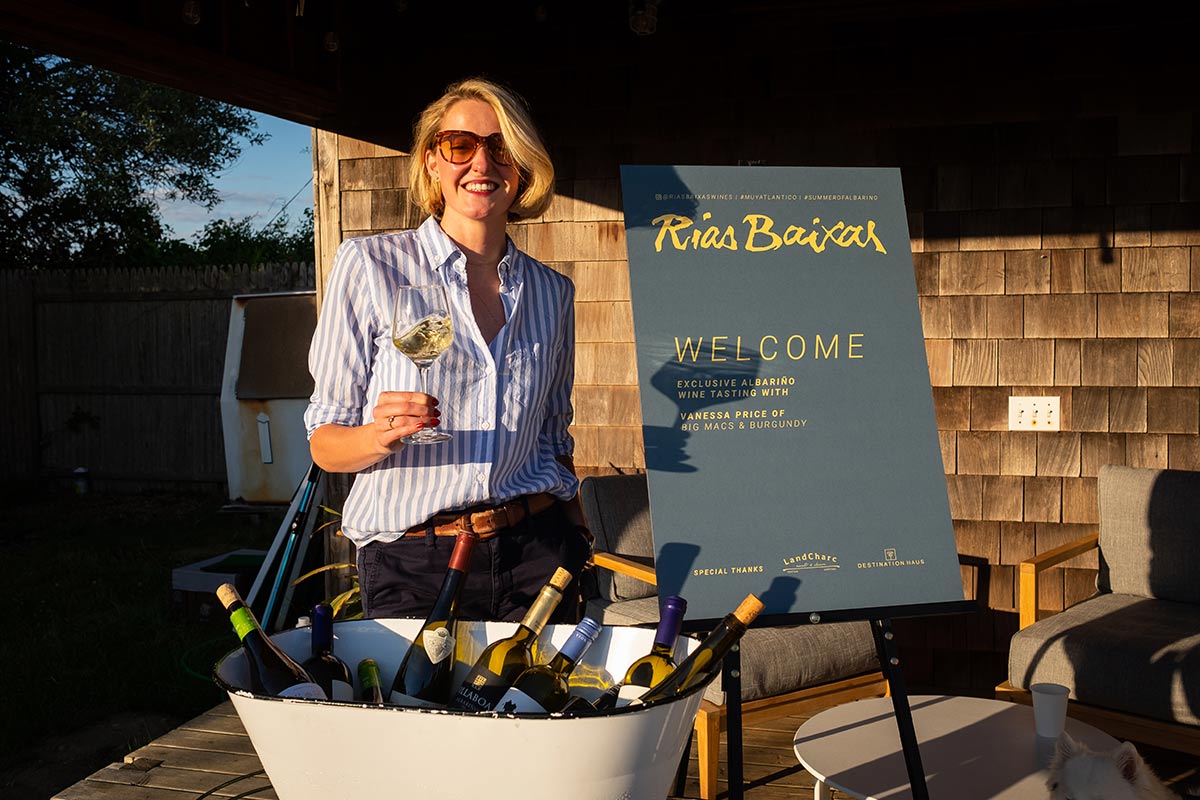 Vanessa Price holding a clear glass of white wine and standing next to a easel sign and bucket of wine bottles at the Boneyard. 