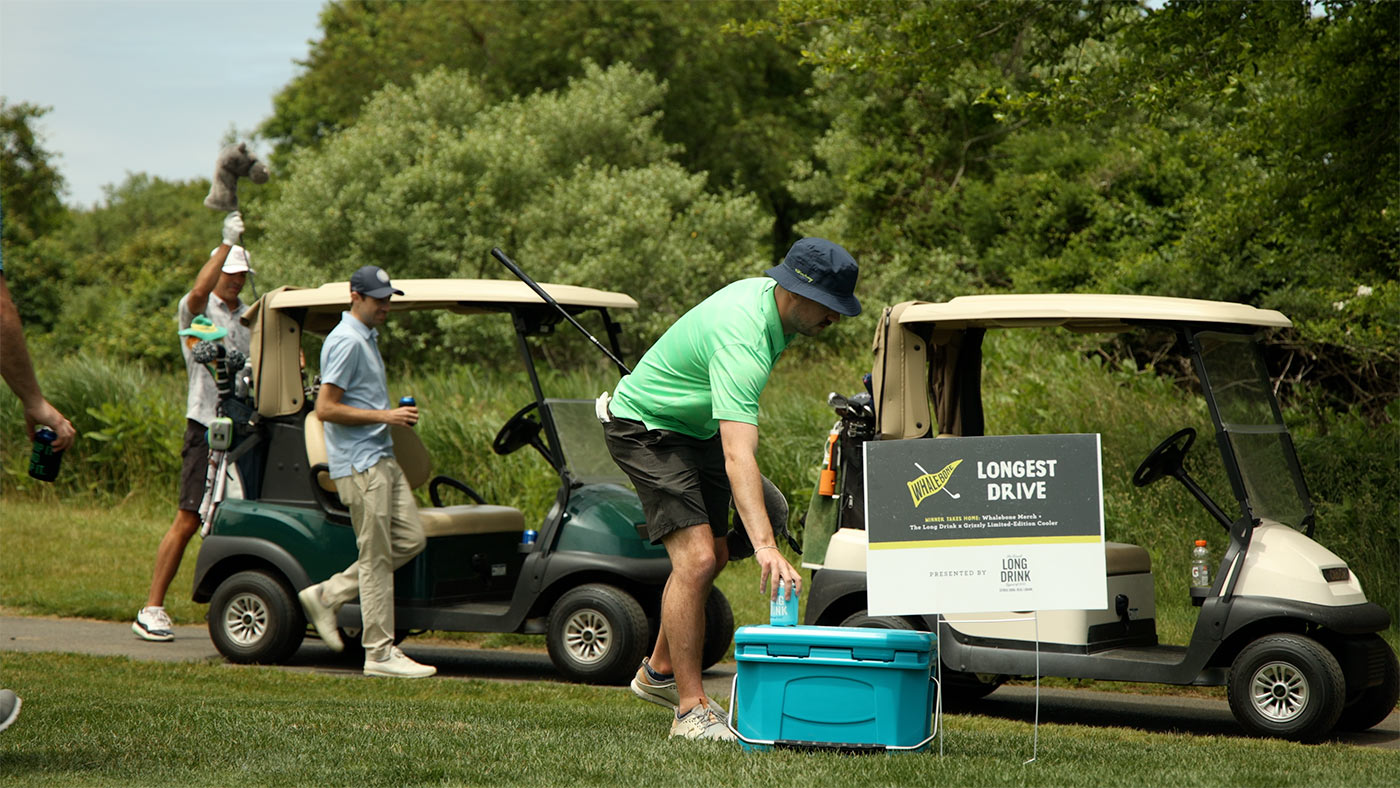 People and parked golf carts. One man in the foreground is reaching for a can that sits atop a cooler