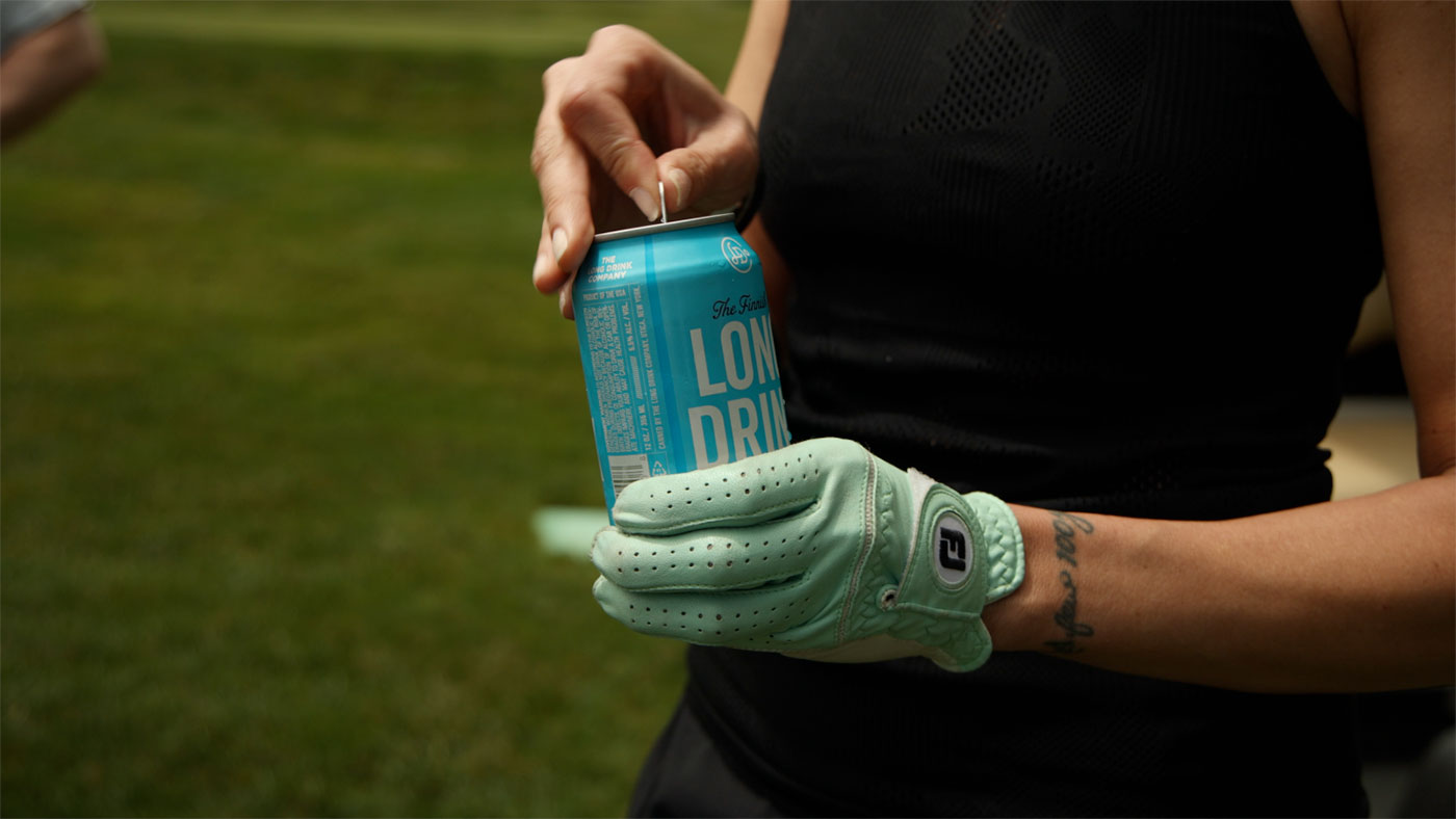 woman wearing a golf glove, holding a long drink can and opening it