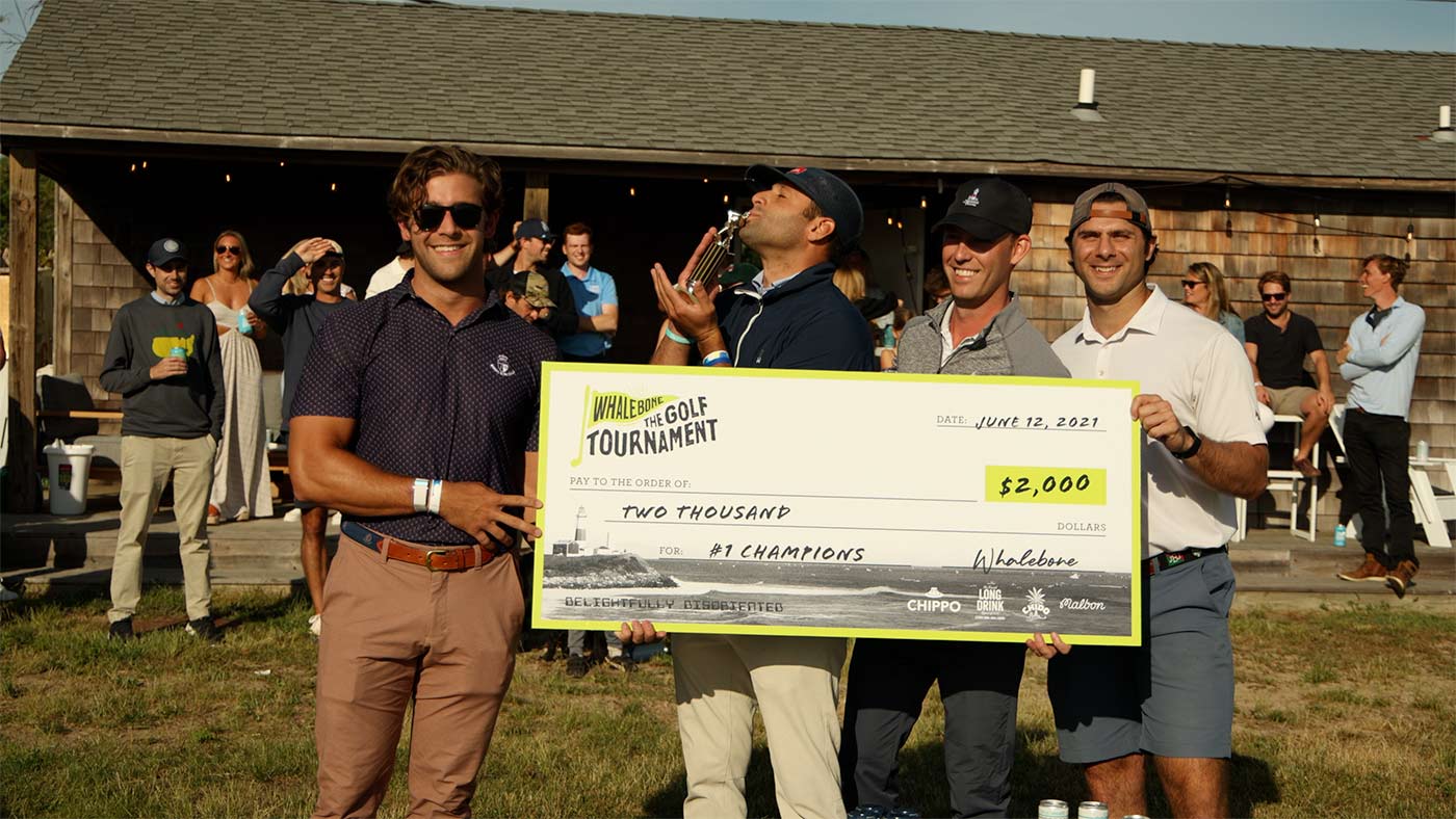 Four men holding a giant check and a trophy. There is a group of people behind them.
