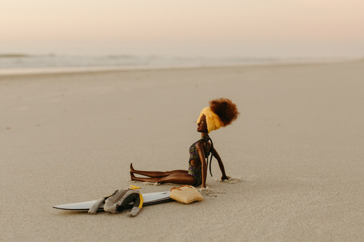 Image of a black barbie doll sitting on the sand with her wet suit, surfboard, and bag.