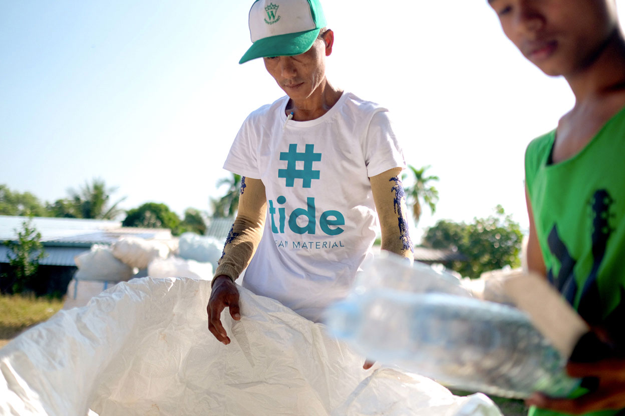 An employee of #tide ocean material holds a white plastic back in the background. In the foreground, a man holds two plastic water bottles. 