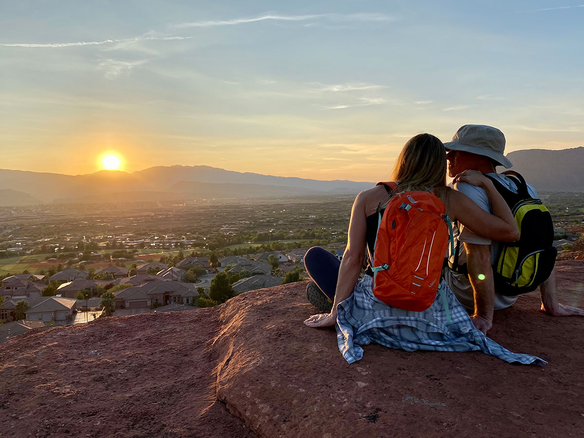 A man and a woman, wearing backpacks, sit close together on a ledge looking over rows of houses. Mountains are in the background with a setting sun.