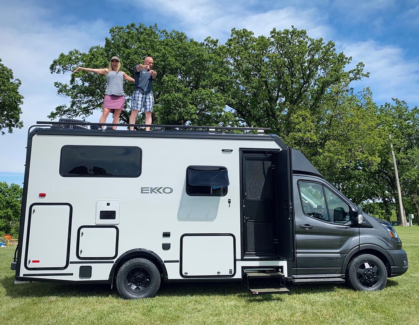 Two adults, a man and a women stand on top of their 2022 Winnebago EKKO. The RV is parked in a grassy field with trees behind it.