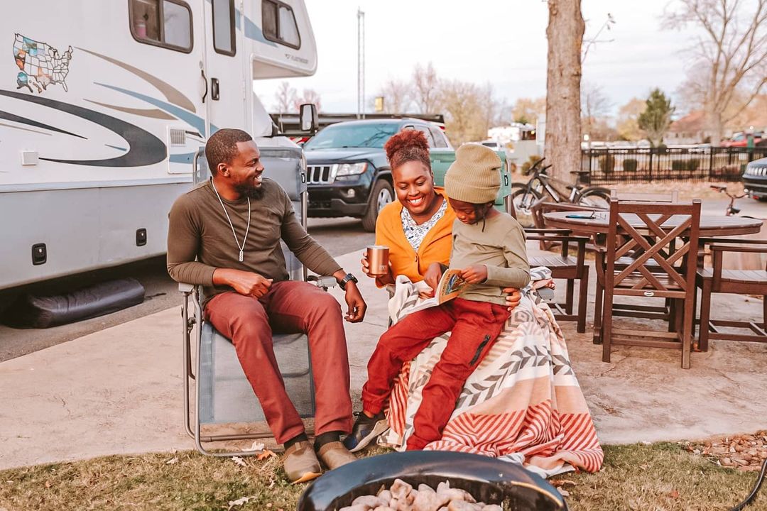 Two adults and a child sit outside of their parked RV in folding chairs. They're dressed for cold weather and sitting in front of a fire pit.