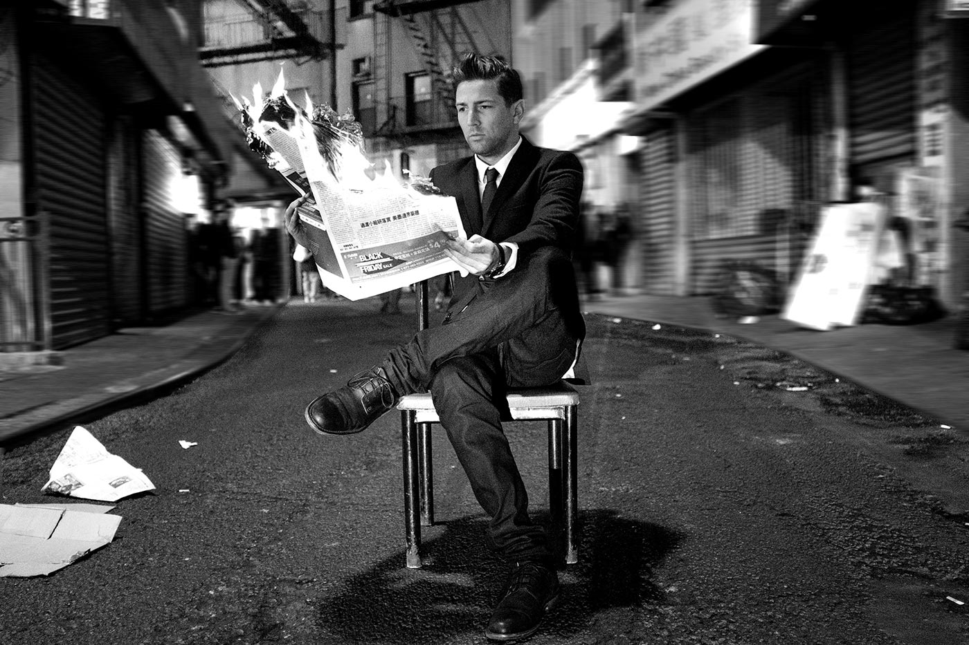 William Grand sitting on a chair in the middle of a city road. He's holding a newspaper that is burning away.