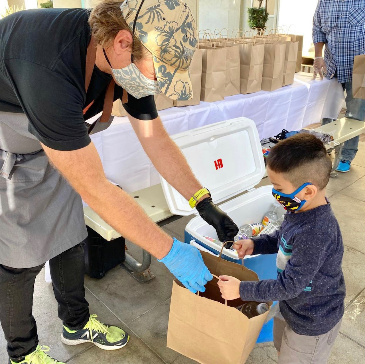 A masked Good Eats volunteer handing a brown paper bag of supplies to a  child