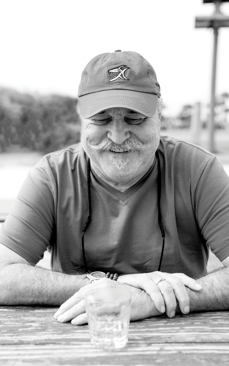 Black and white photograph by Gunner Hughes of John Miglis. It's an older man smiling with his eyes closed. He's wearing a hat and tshirt. 