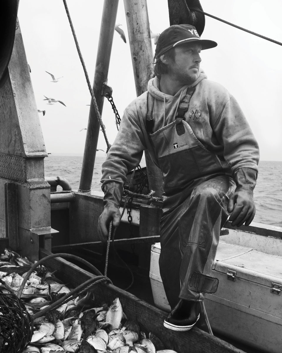 Black and white photograph by Max Foley or Tyler Maguire. A man is sitting on the edge of a boat, dressed in fishing gear, with his foot raised and rested on a bin of fish.