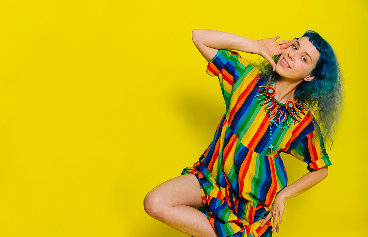 Jess Joy leaning to the right. She's dressed in a dress of rainbow colors and stands in front of a blank yellow background. 