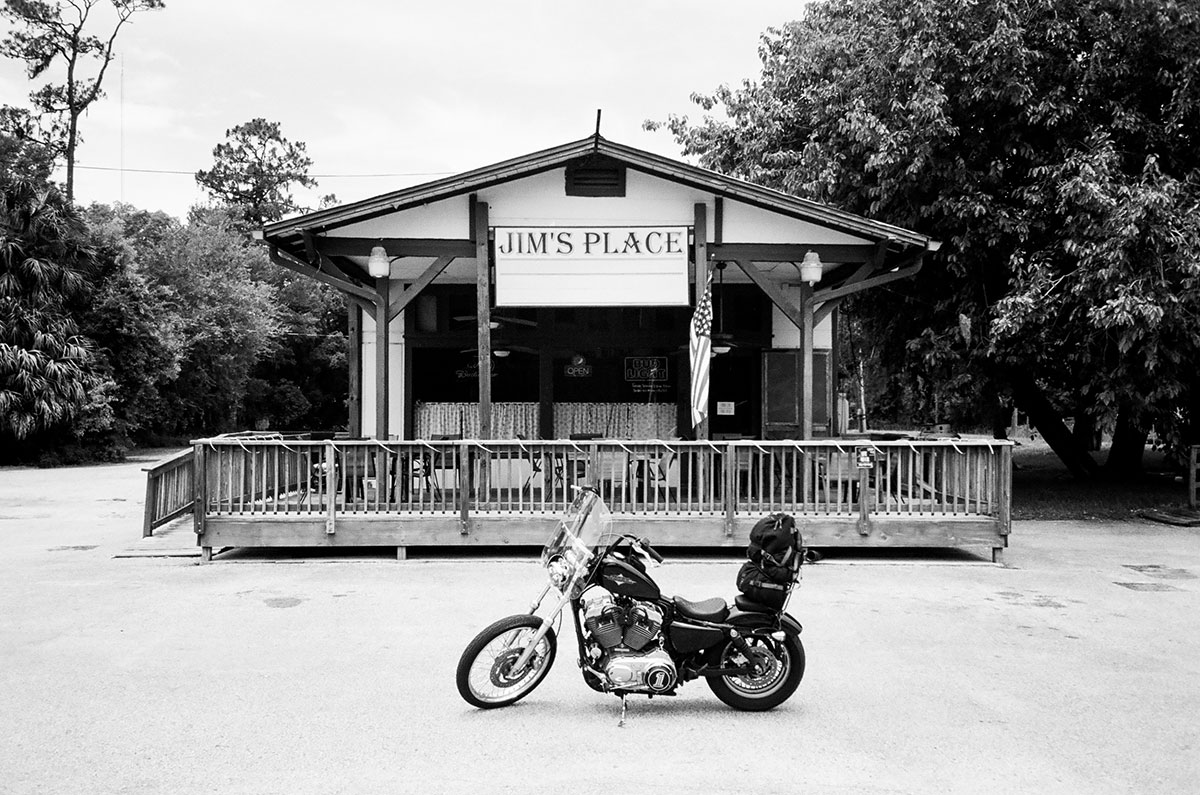 Black and white photograph by Gunner Hughes of Jim's Place, a small bar or restaurant building with a wraparound wood porch. In front of the building sits a riderless motorcycle in the parking lot.