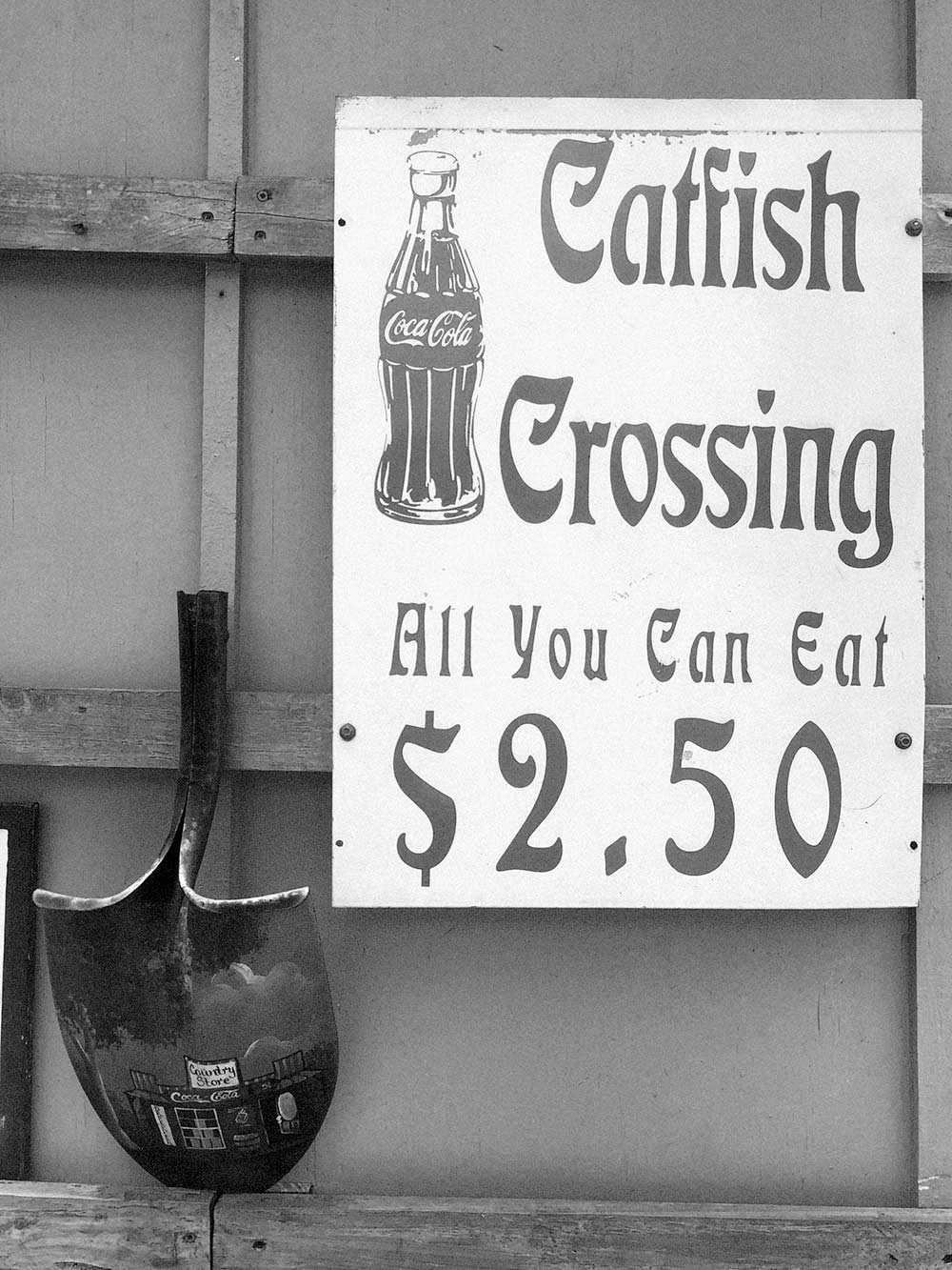 black and white photograph by Gunner Hughes of a sign that reads catfish crossing, all you can eat $2.50