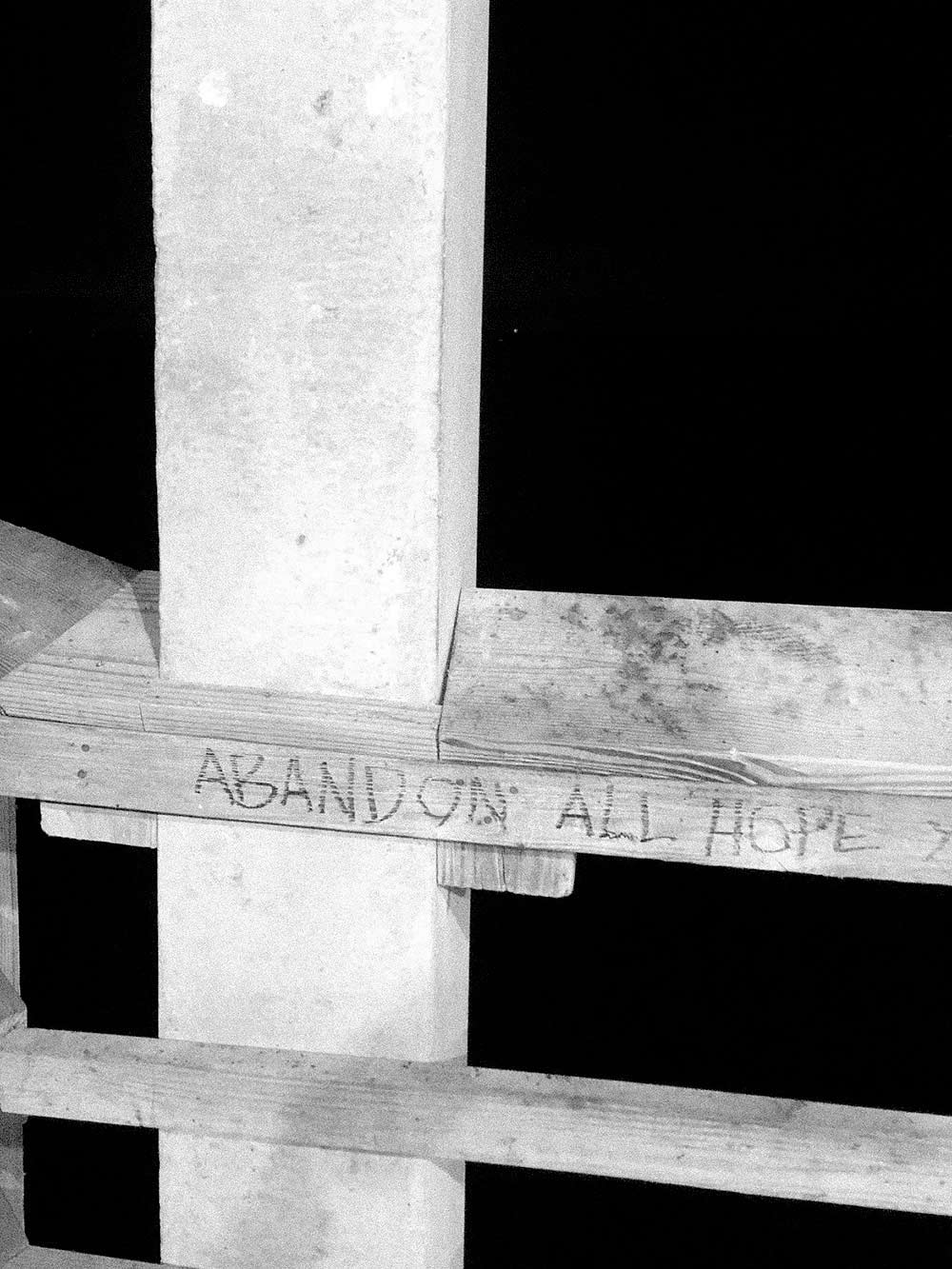 black and white photograph by Gunner Hughes of a wood deck. Scratched into it are the words abandon all hope.