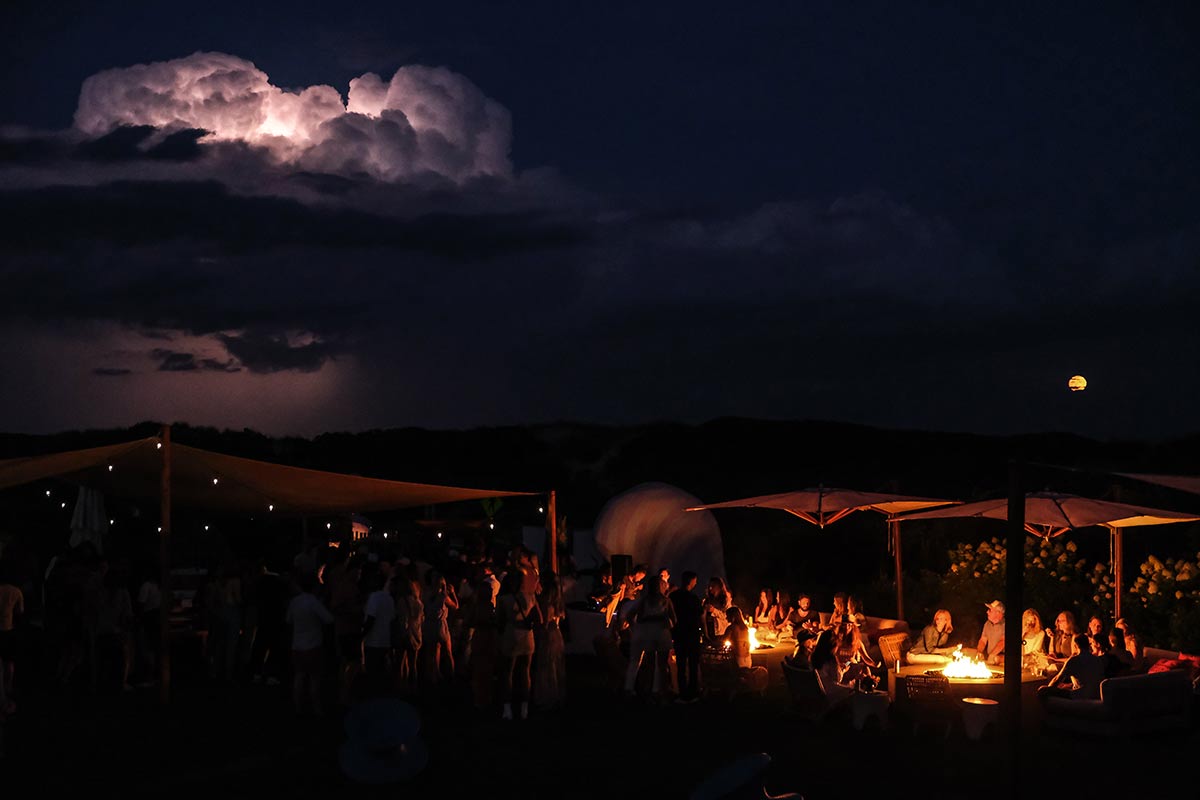 A wide shot with fires and guests in the forefront and a lightening thunderstorm in the background at the Tanqueray event at Hero Beach Club in Montauk 