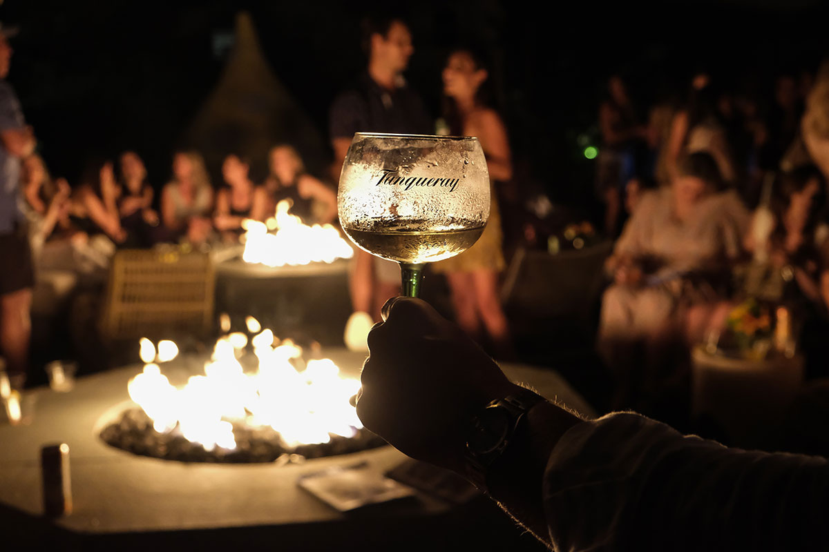 A hand holds a glass in front of a backdrop of fires and guests at the Tanqueray event at Hero Beach Club in Montauk 