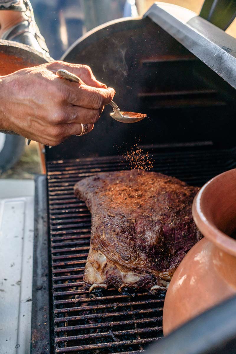 A man holding a spoon sprinkles seasoning over ribs on a grill.