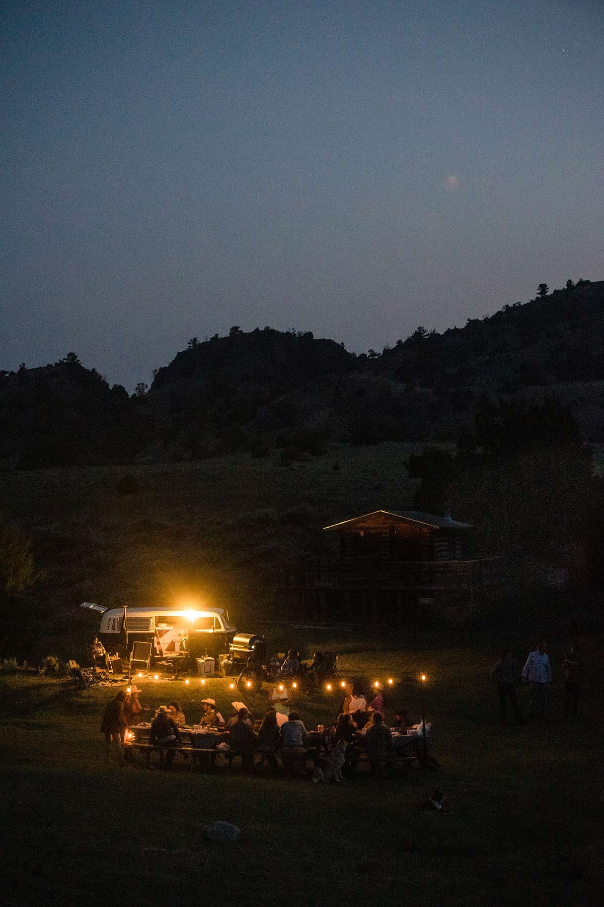 Photograph of late evening. A let vw bus sits in the background. In the foreground people sit together under a lit table.