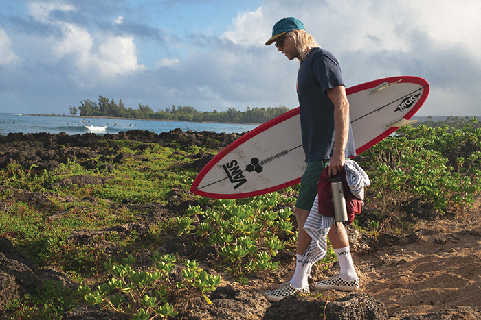 Dane Gudauskas walking toward the water, holding a surf board and wearing Vans Eco Theory Collection.