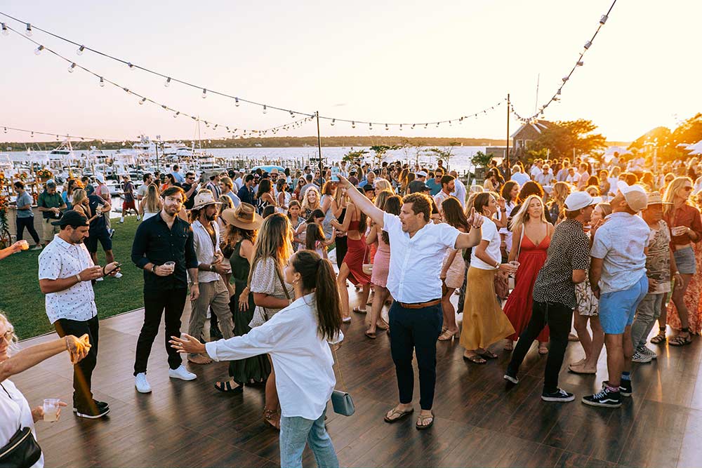 A crowd of people moving on a dance floor at the Whalebone Magazine Sixth Anniversary party at the Montauk Lake House. A marina full of boats is in the background.