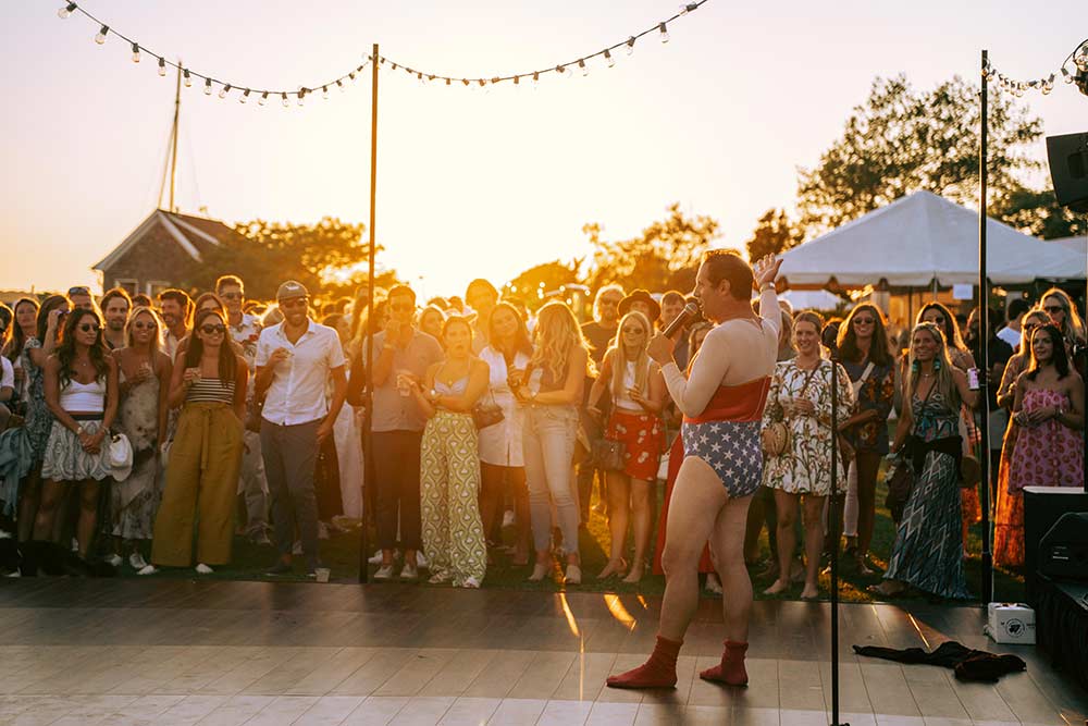 Comedian Seth Herzog dressed in a wonder woman costume holds a mic and speaks to a crowd of smiling people at the Whalebone Magazine Sixth Anniversary party at the Montauk Lake House