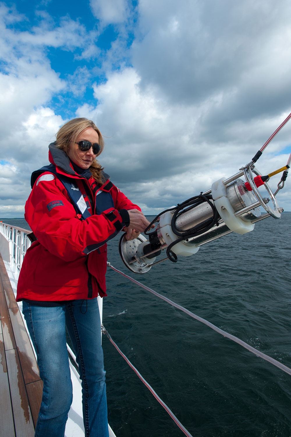 Alexandra Cousteau hauling on boat deck. She's wearing a red jacket, sunglasses, and jeans. 