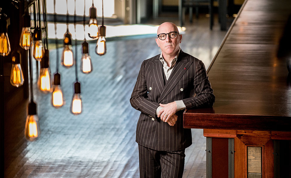 Maynard James Keenan dressed in a pin stripe suit leans against an empty long wooden counter.