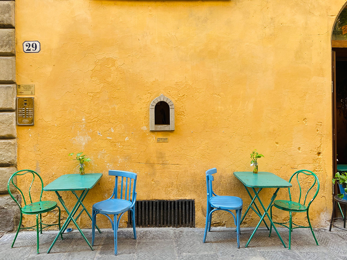In a yellow wall beset by colorful chairs and tables is a wine window in Florence, Italy