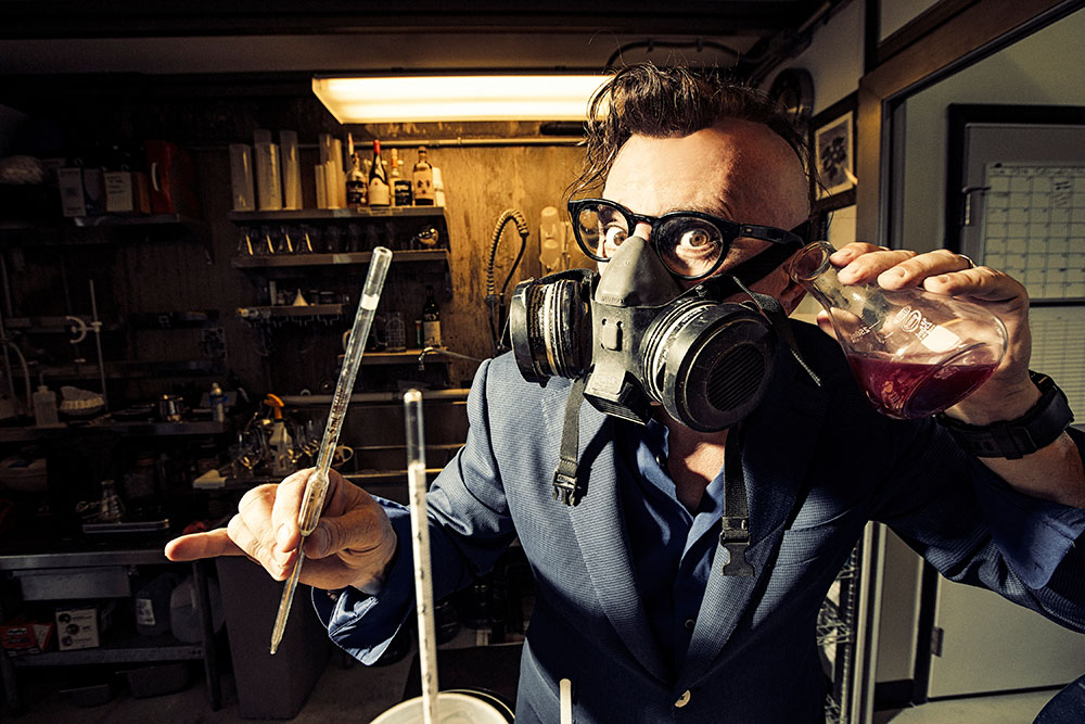 Maynard James Keenan wearing a gas mask holds a glass flask and a volumetric pipettes in a lab. 