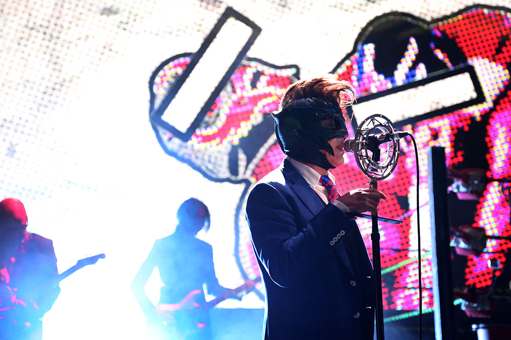 Maynard James Keenan performs wearing a mask and standing in front of a microphone. He wears a blue suit and tie. Behind him is a wall of lights and two people holding guitars.