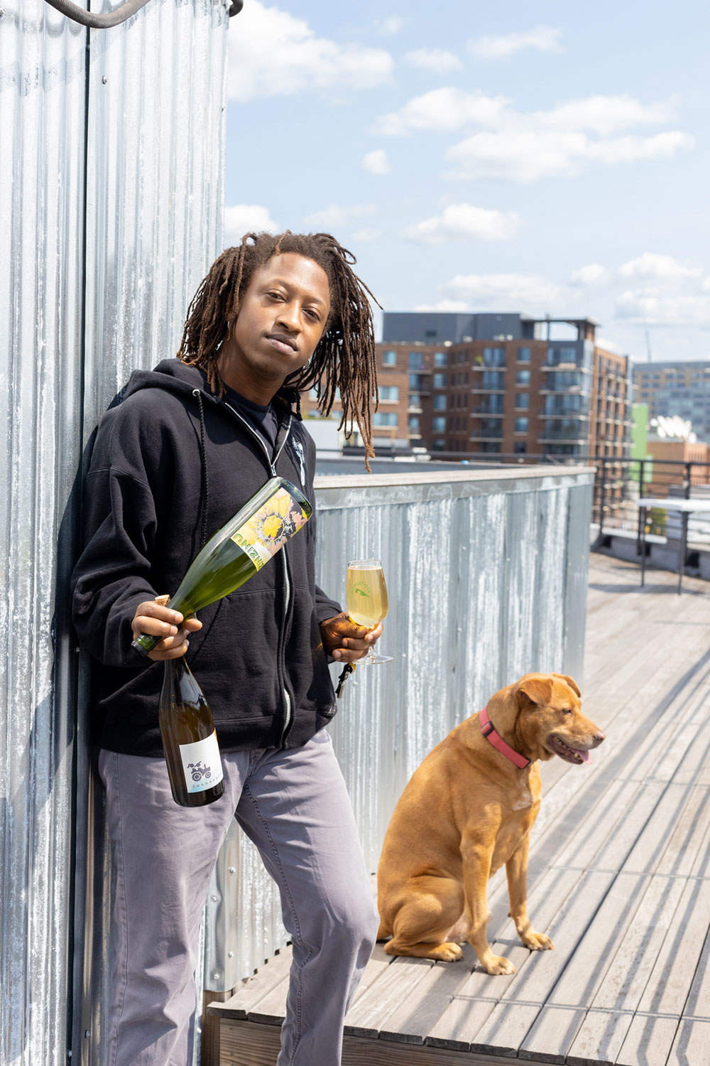 Eric Moorer holding wine bottles and a wine glass. He stands against an industrial background. A sitting dog is behind him.