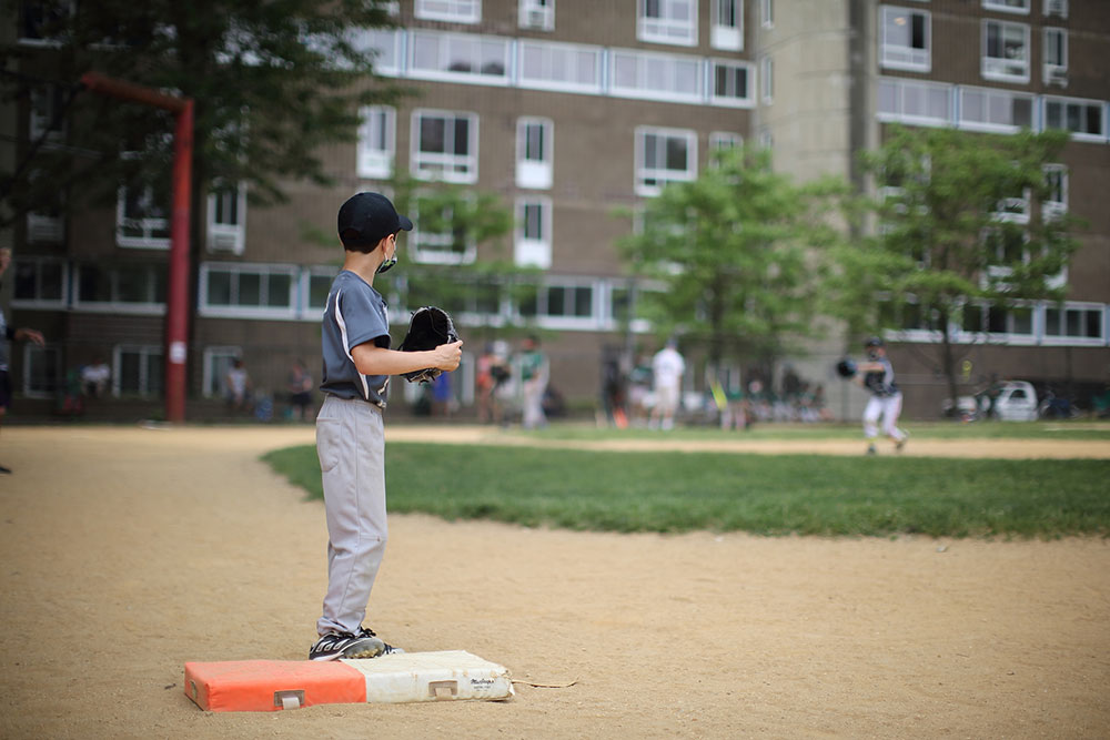 Laura June Kirsh's nephew playing little league baseball. A young child stands on the catches mat. he holds a glove and wears a cap. In the background there is the larger baseball field and apartment buildings.