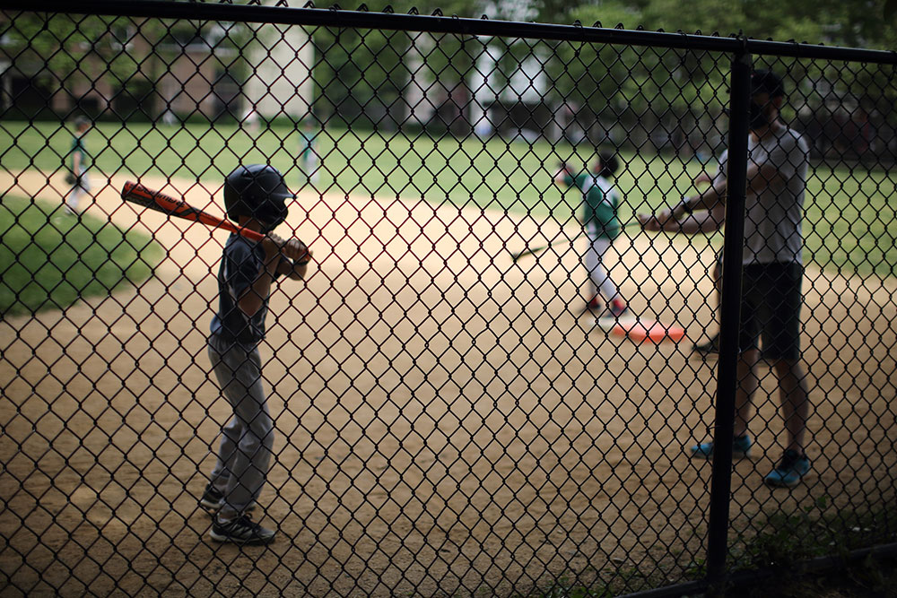 Laura June Kirsh's nephew playing little league baseball. He wears a batter's helmet and is holding a bat preparing to swing. In the foreground is a black chain fence.