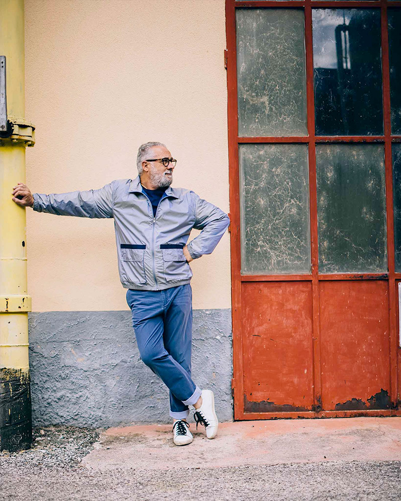 An older man with glasses stands in front of a light pink wall. He leans on a yellow pipe. He's wearing Rhone Nanoprojects Bowie Windbreaker in Ashley Blue