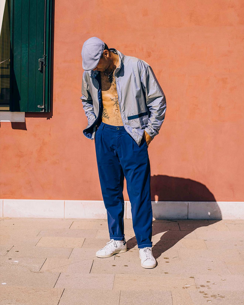 A man in a light jacket and cap wears noprojects Enzo Trouser in Indigo Blue and stands outside in the sun against a burnt orange wall