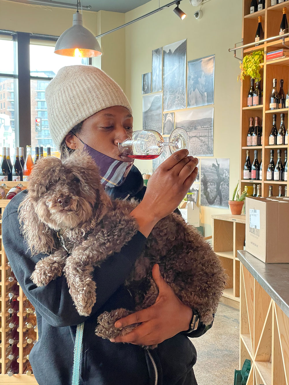 Eric Moorer drinks red wine out of a glass while he holds a small furry brown dog. Eric is standing in a store surrounded by shelves of wine.
