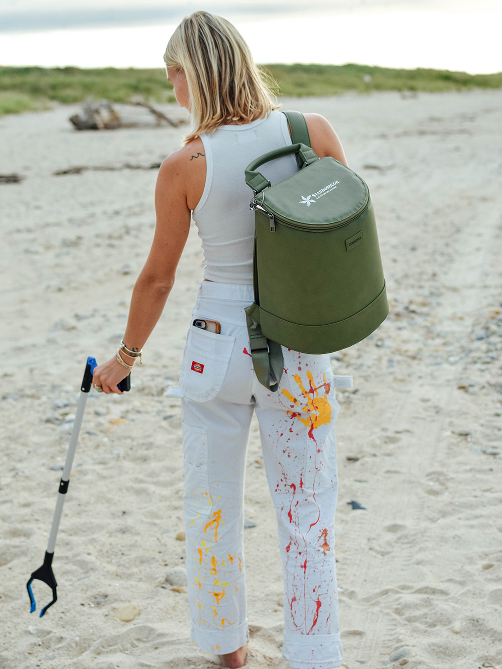a woman wears a green back pack and picks up trash on gin beach in montauk, new york