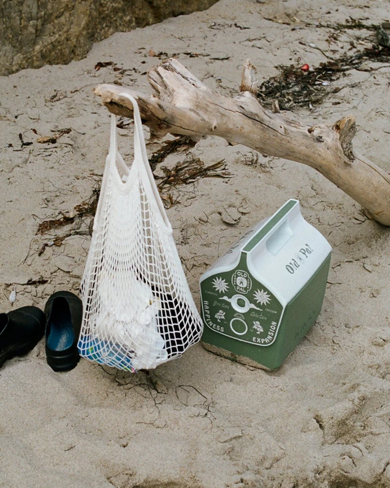 A beach scene with black shoes, empty net bag and a sage green Old Pal Provisions x Igloo Eco Cool “Cold Pal”,