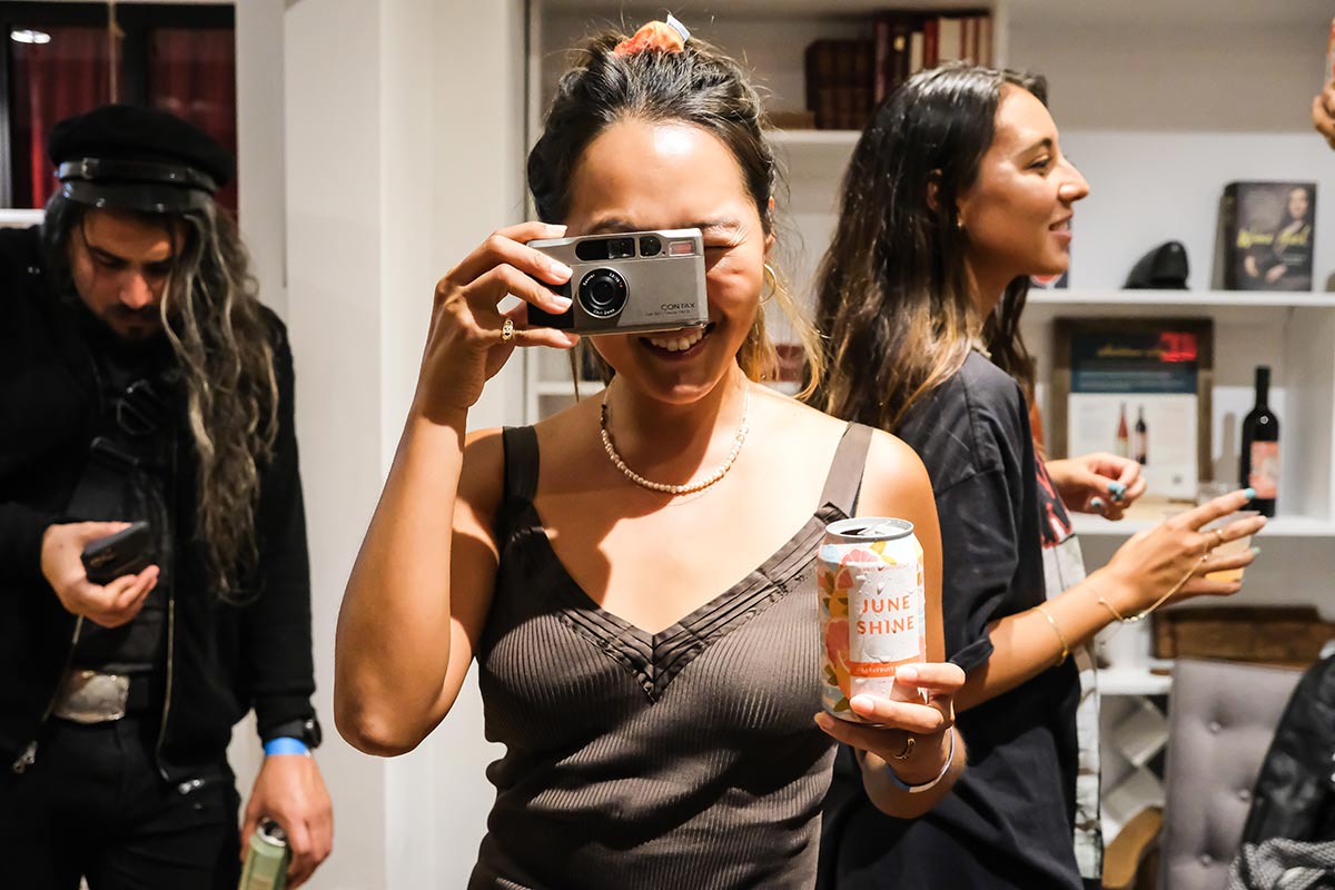 A woman holds a can of JuneShine in one hand and a camera to her face with the other at the Governors Ball pre-game bash with JuneShine and Whalebone Magazine on Bleecker Street.