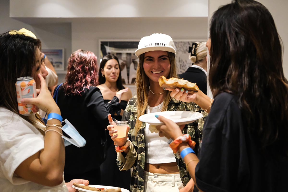 A group of people stand in a lit room. In the foreground three women are drinking and eating pizza at the Governors Ball pre-game bash with JuneShine and Whalebone Magazine on Bleecker Street.