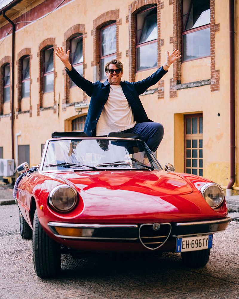 A man wearing glsases, a white tshirt and wool jacket holds his hands in the air as he stands in a red sports car