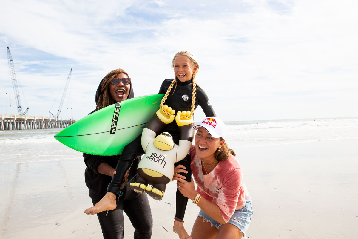 Tyko the Grom Ripper being held up by Gigi Lucas and Carissa Moore with giant Sun Bum chain