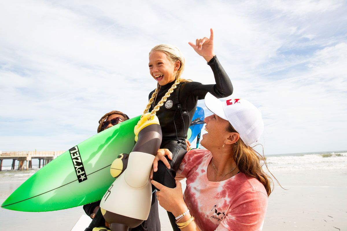 Tyko the Grom Ripper being held up by Gigi Lucas and Carissa Moore with giant Sun Bum chain and shaka sign