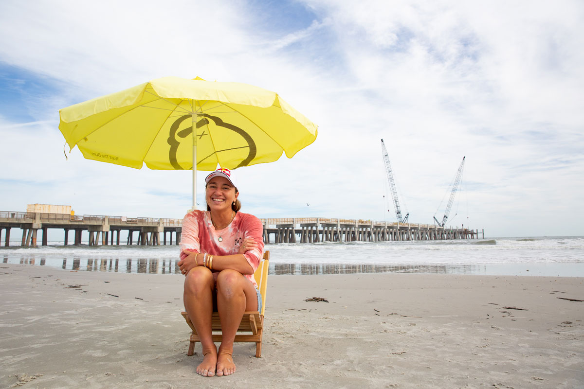 Carissa Moore sitting on beach under Sun Bum umbrella with pier in the background.