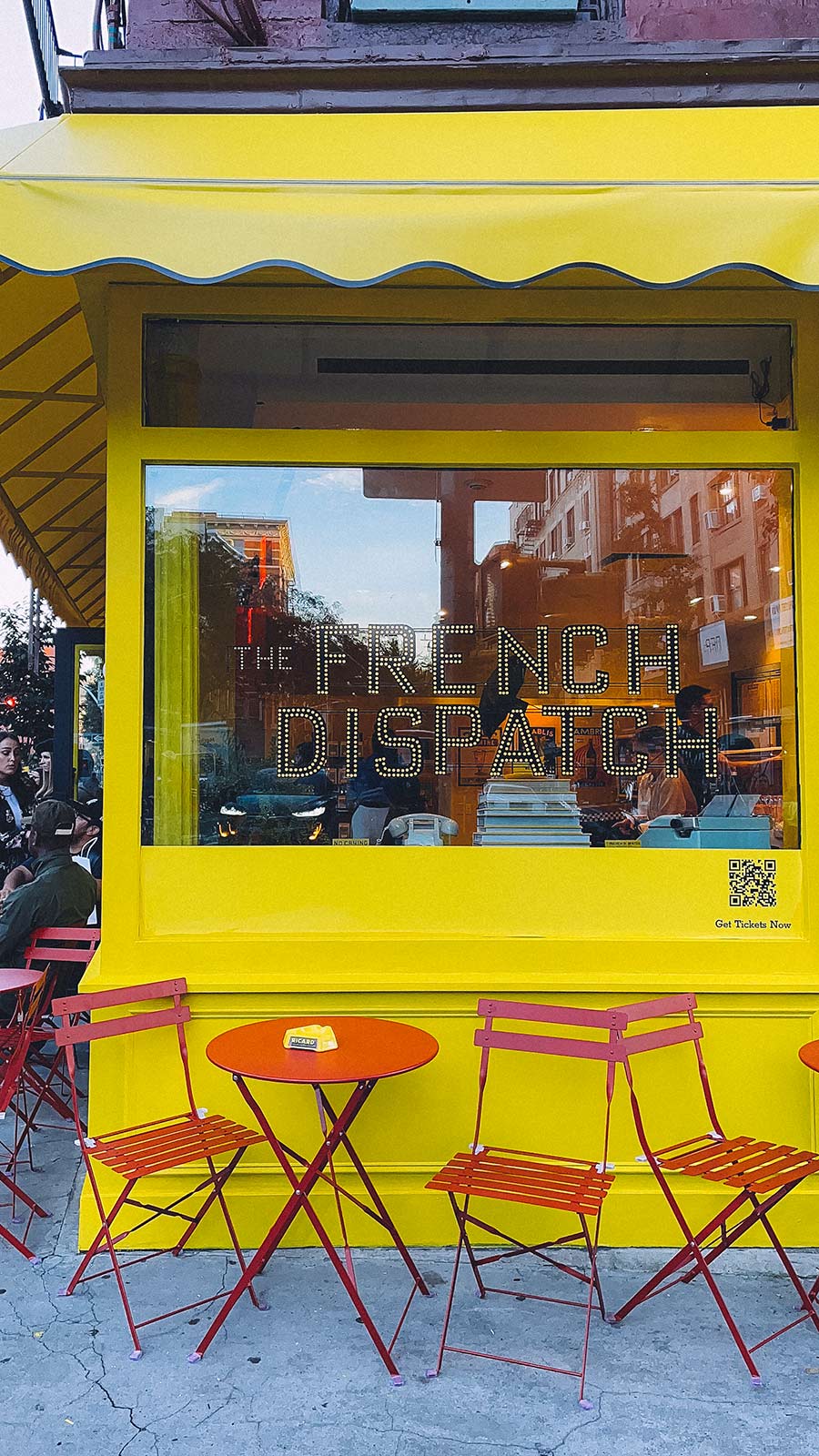 The corner of a yellow building with a large window. Cafe tables and chairs sit against the walls at Le Sans Blague, the The French Dispatch takeover of Whalebone on Bleecker