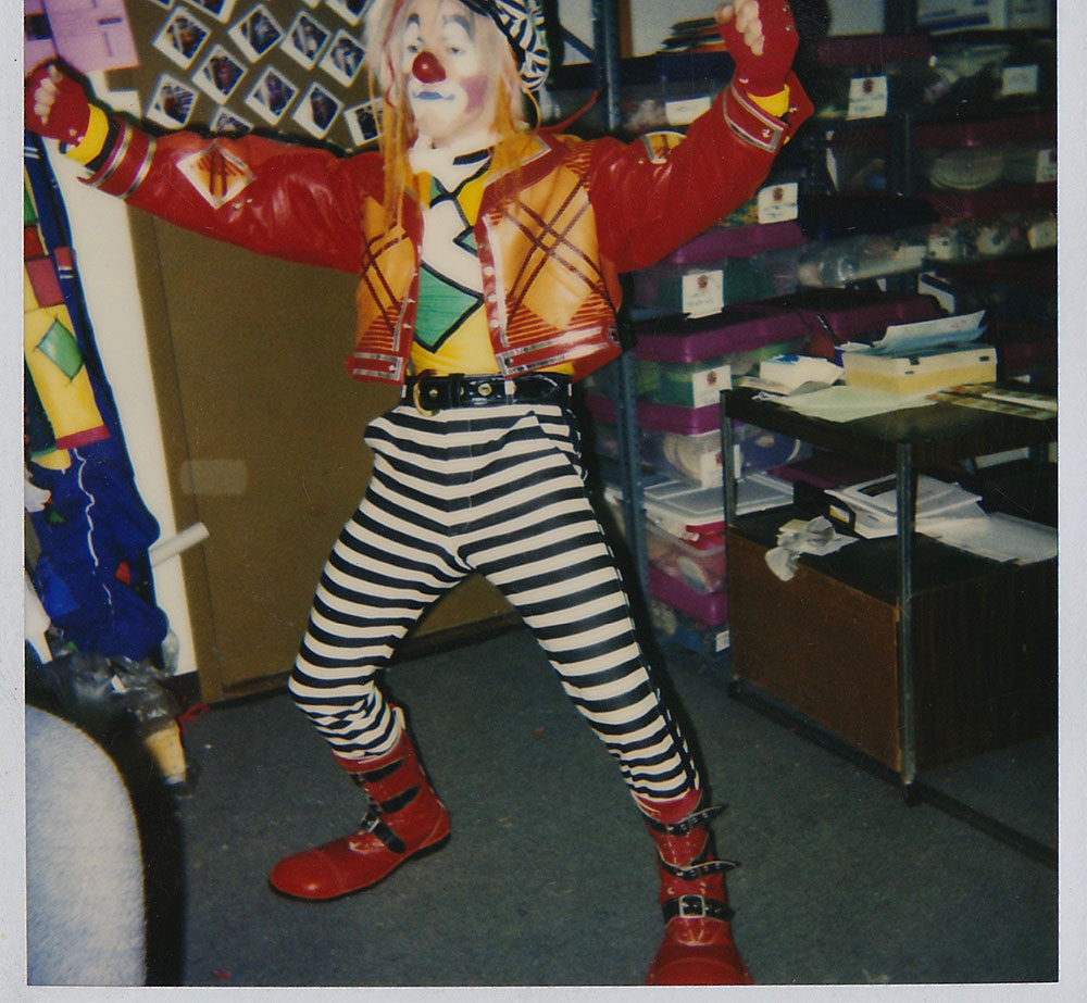 Steve-o in full clown attire. A man in striped pants, red shoes, yellow and green shirt, and red leather jacket poses in a storage room. His face is painted in clown make up and he's wearing a wig. 
