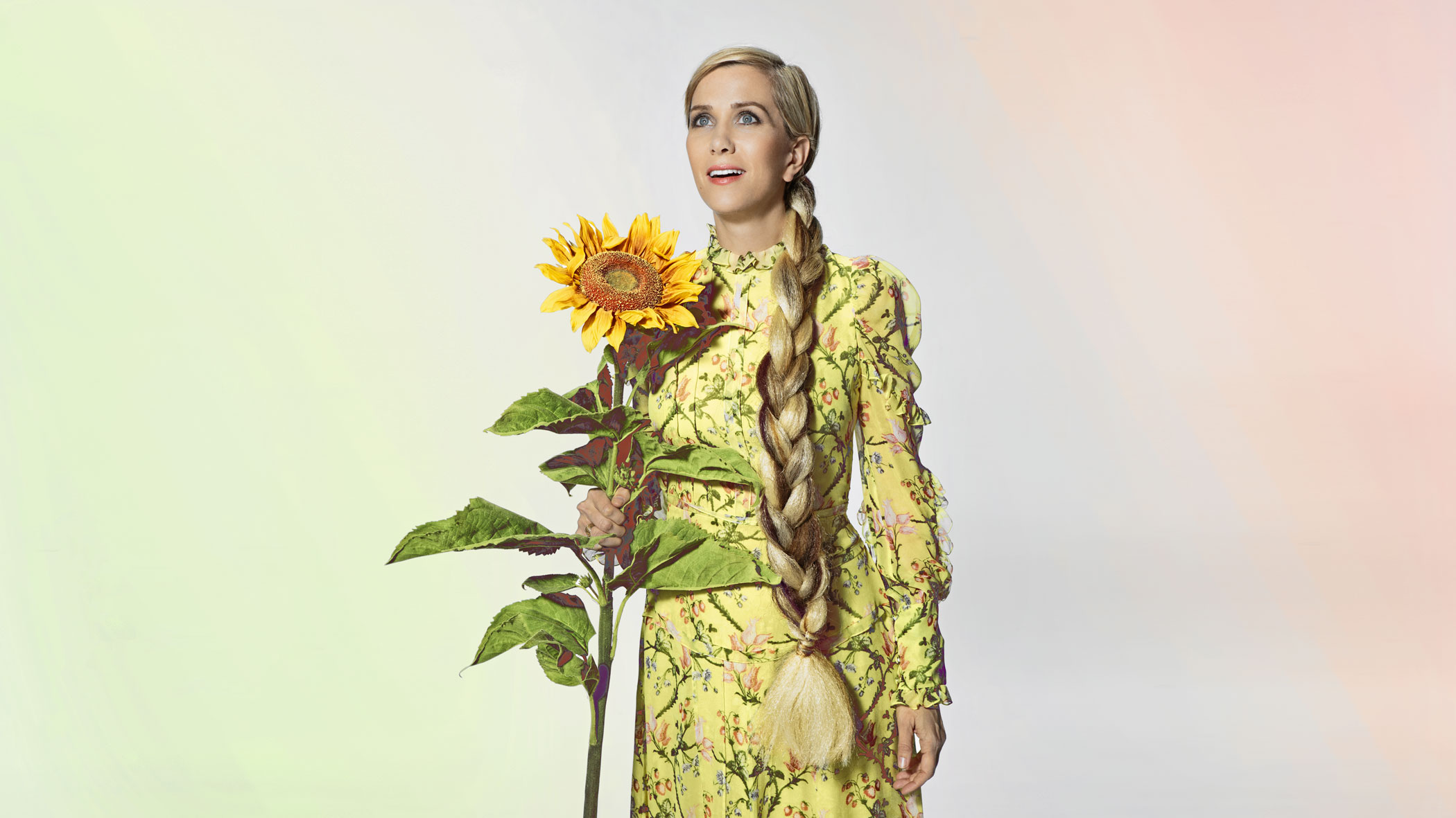 Kristen Wiig with big sunflower and big braid and wearing a yellow dress with flower print.