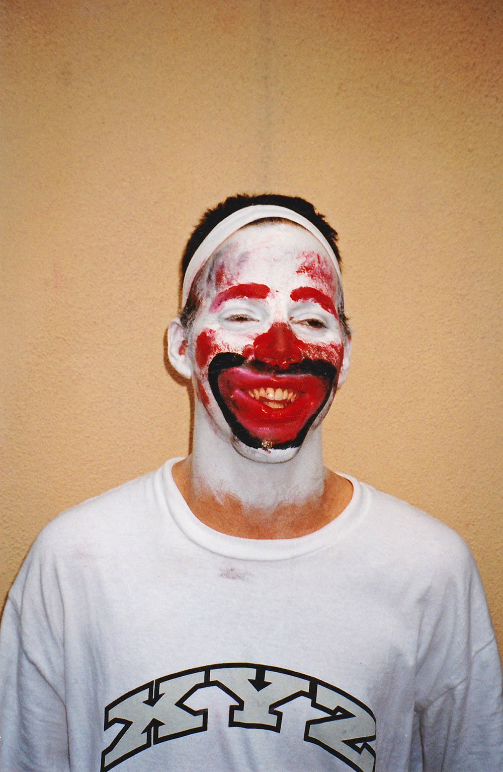 picture of Steve-O's first attempt at clown makeup. A man wearing a white tshirt with the letters XYZ has white, red, and black paint all over his face. 