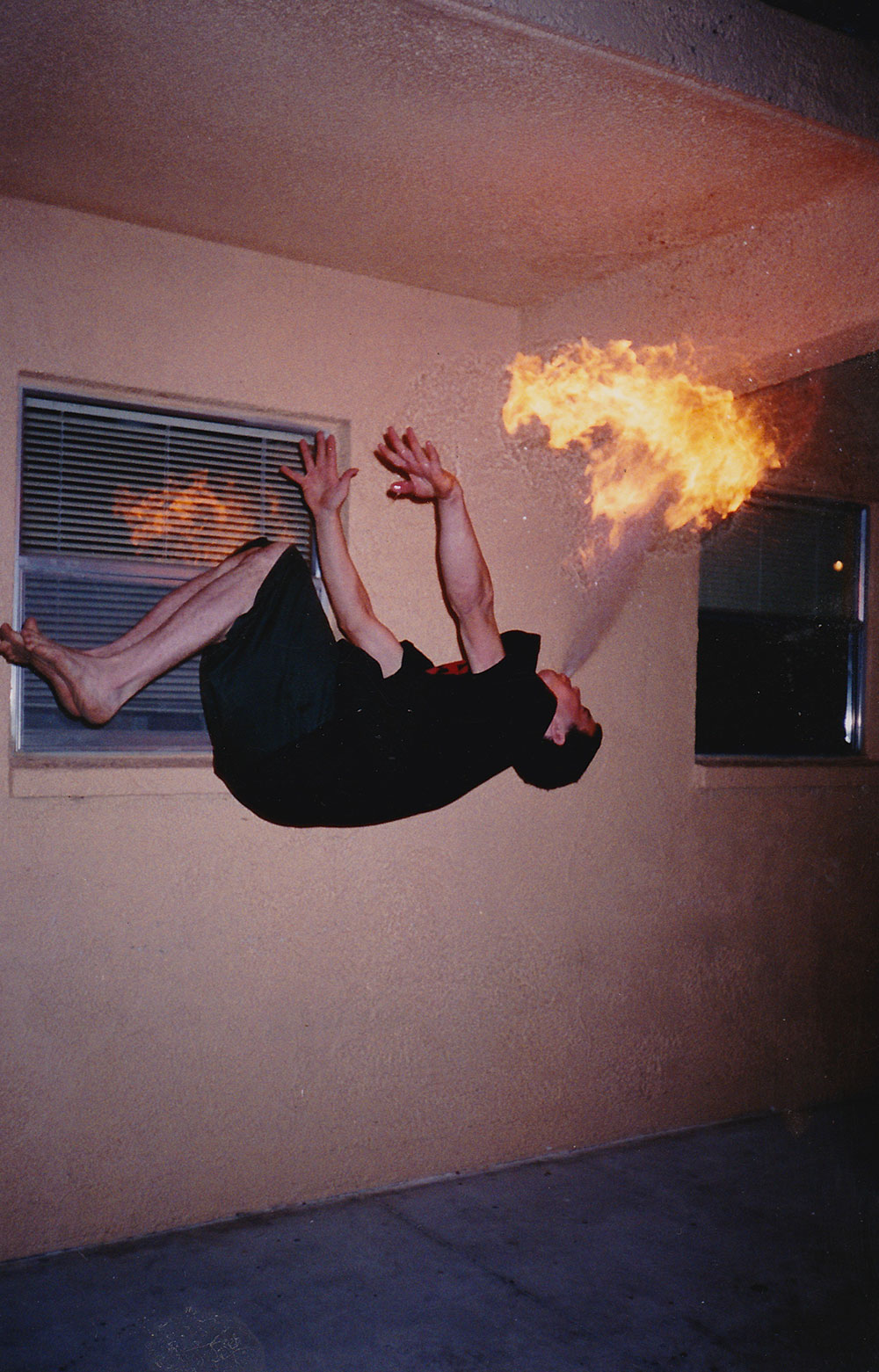 Steve-O doing a backflip while breathing fire. A man wearing black shorts and shirts is blowing fire out of his mouth while halfway through a back flip.