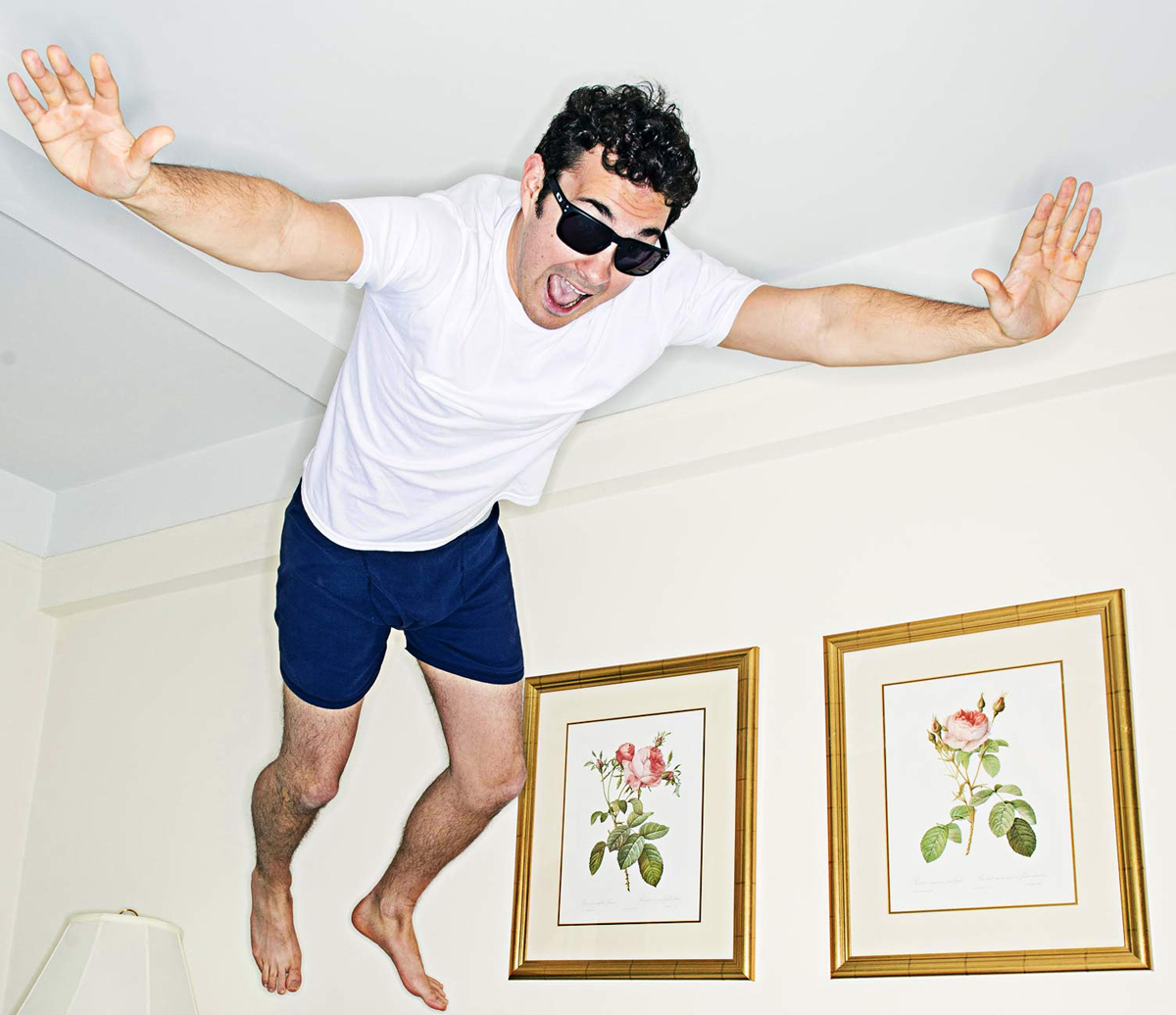 Mark Normand jumping onto bed in the air. He wears sunglasses and a white under shirt and blue shirts. Two prints of roses hanging on the beige wall behind him. 
